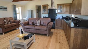 A spacious living room with a brown leather sofa and a wooden coffee table, a kitchen with wooden cabinets and modern appliances, and a high ceiling with exposed beams. - Glebe Cottage (Alnwick)