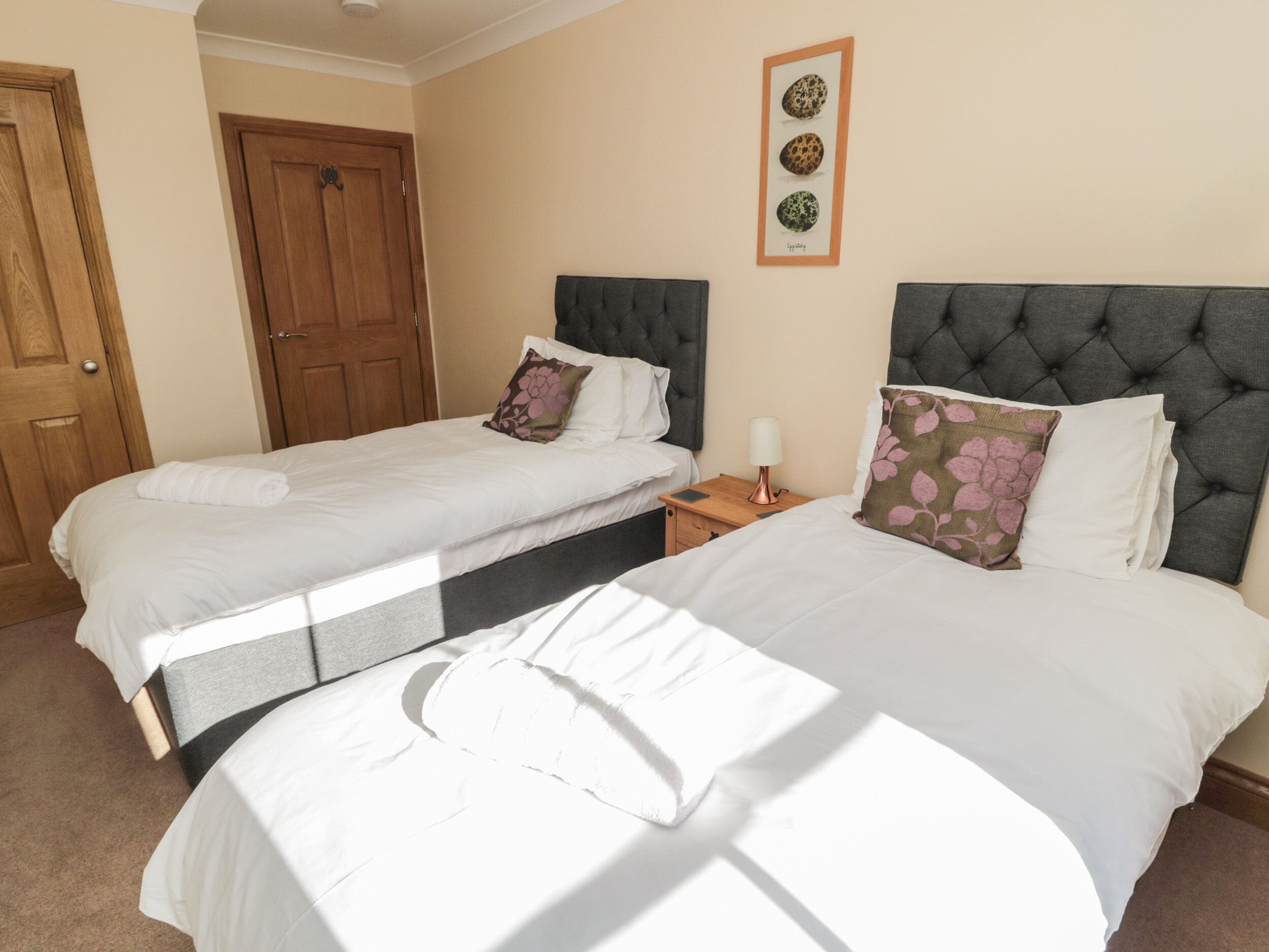 A hotel room with two beds, a wooden door, a framed picture on the wall, and a nightstand with a lamp.