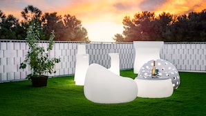 Un área de estar al aire libre con muebles blancos modernos y una planta en maceta, frente a una cerca blanca y árboles durante el atardecer.
