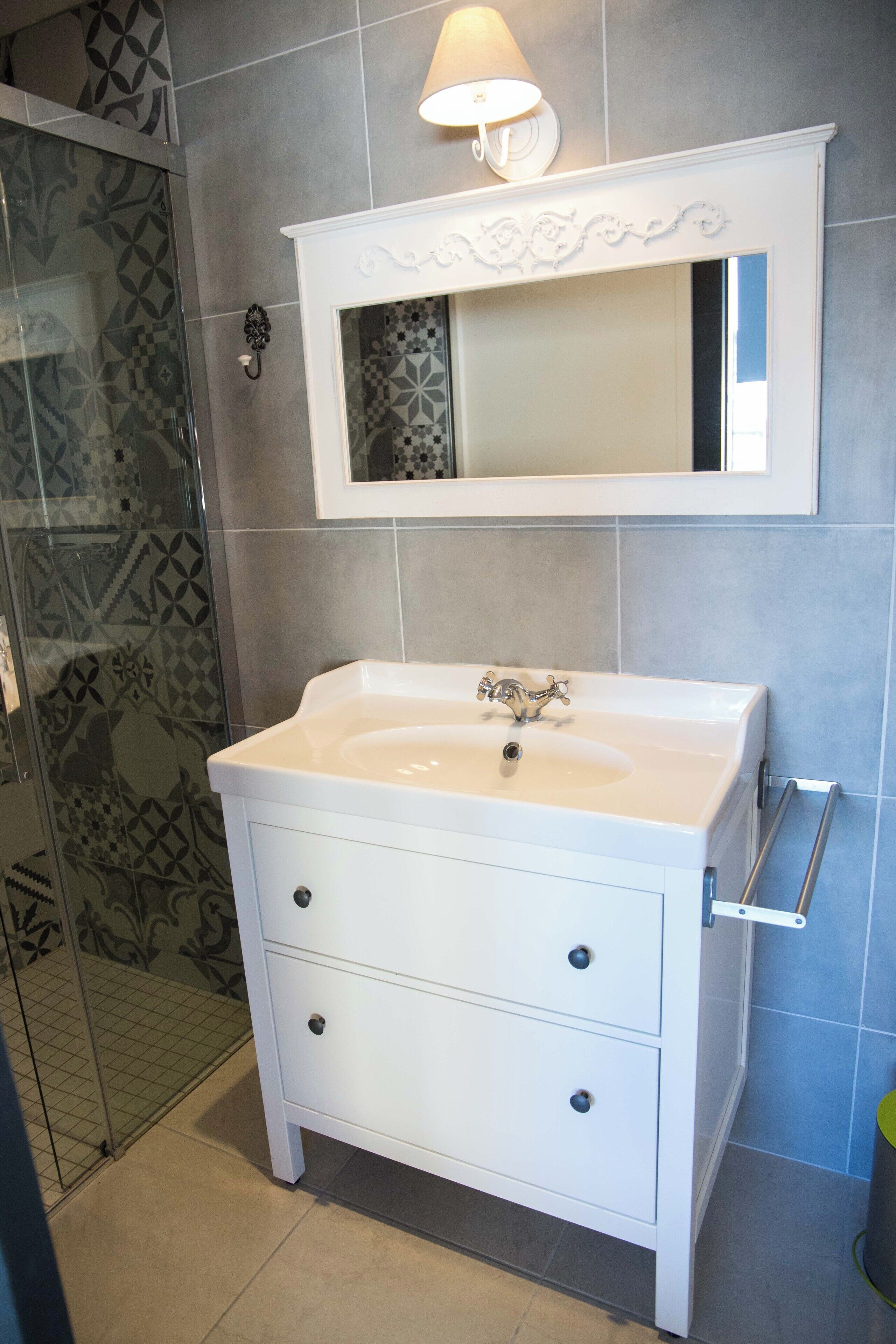 A bathroom with a white vanity, a mirror with decorative molding, and a shower with patterned tiles.