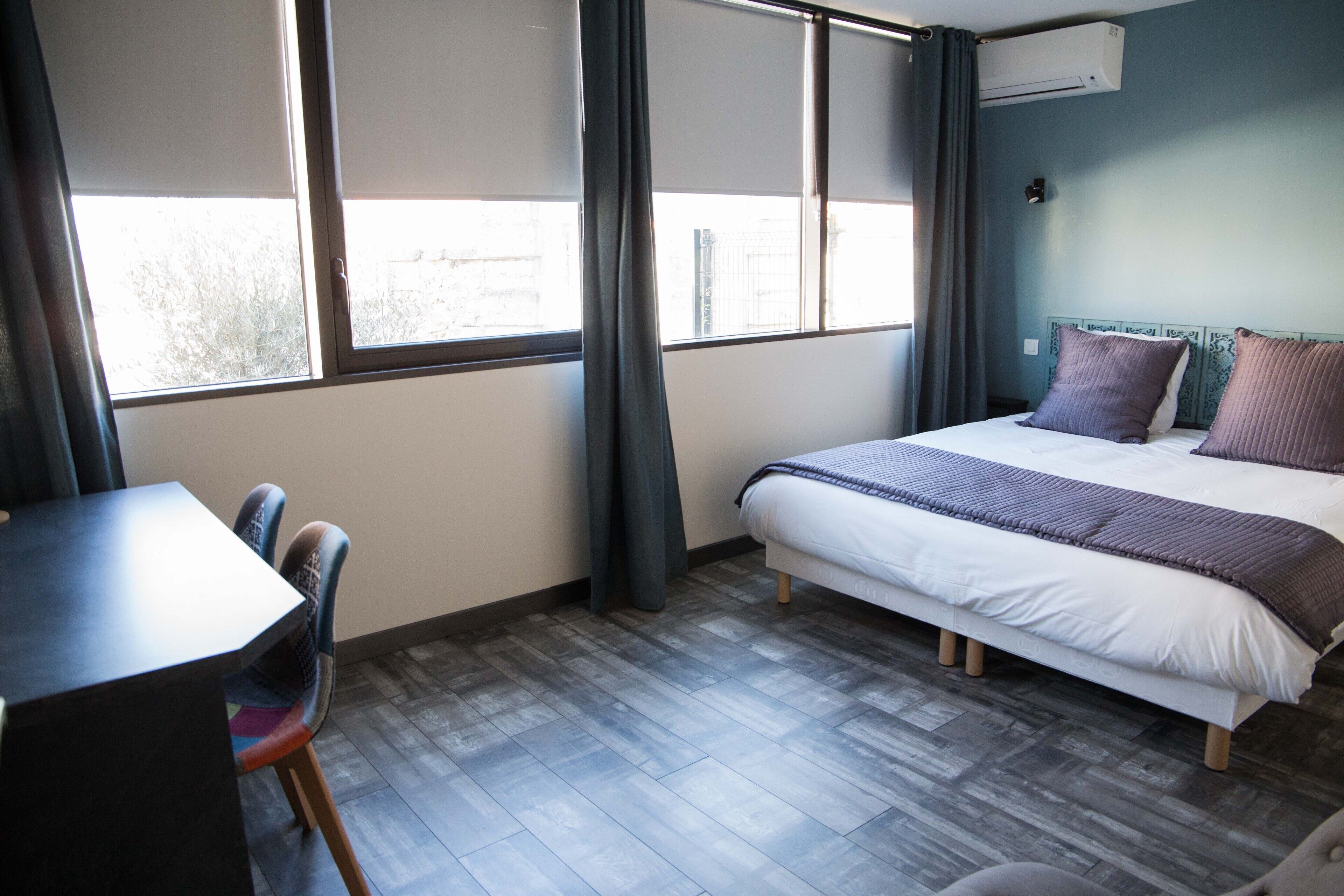 Hotelkamer met een bed, een bureau en een stoel. Er is een raam met gordijnen, een airconditioning aan de muur en uitzicht op buiten.