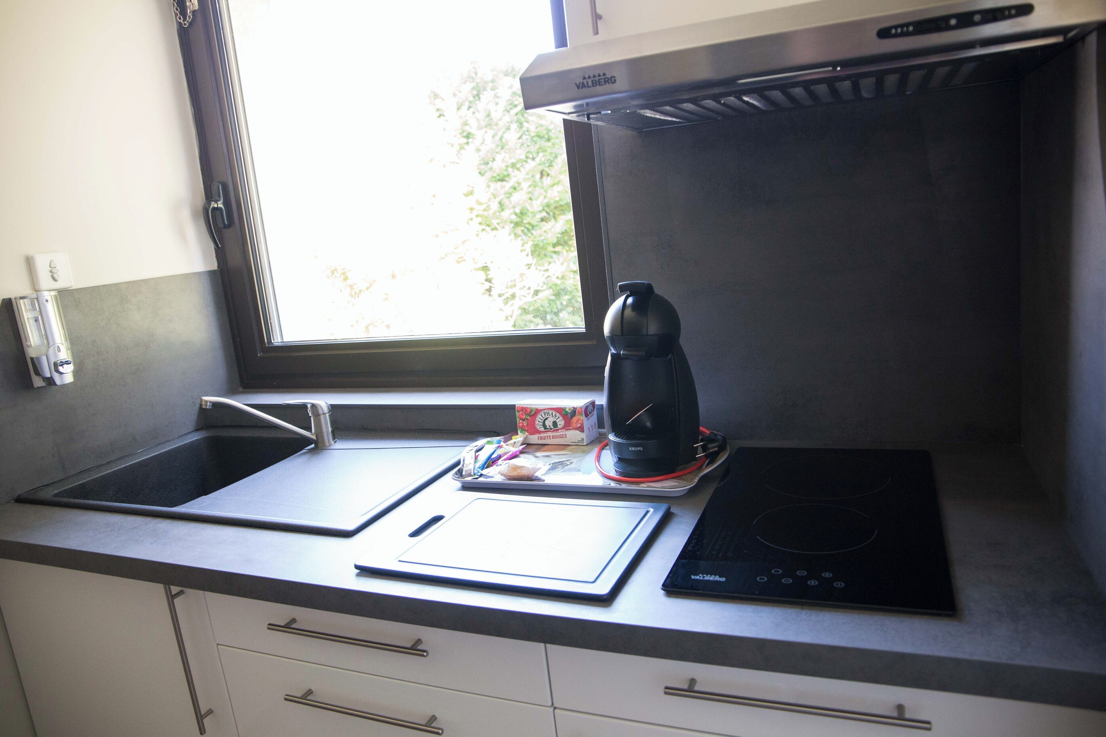 Une cuisine avec une fenêtre, une machine à café, une boîte de capsules de café et une tablette posée sur le comptoir.