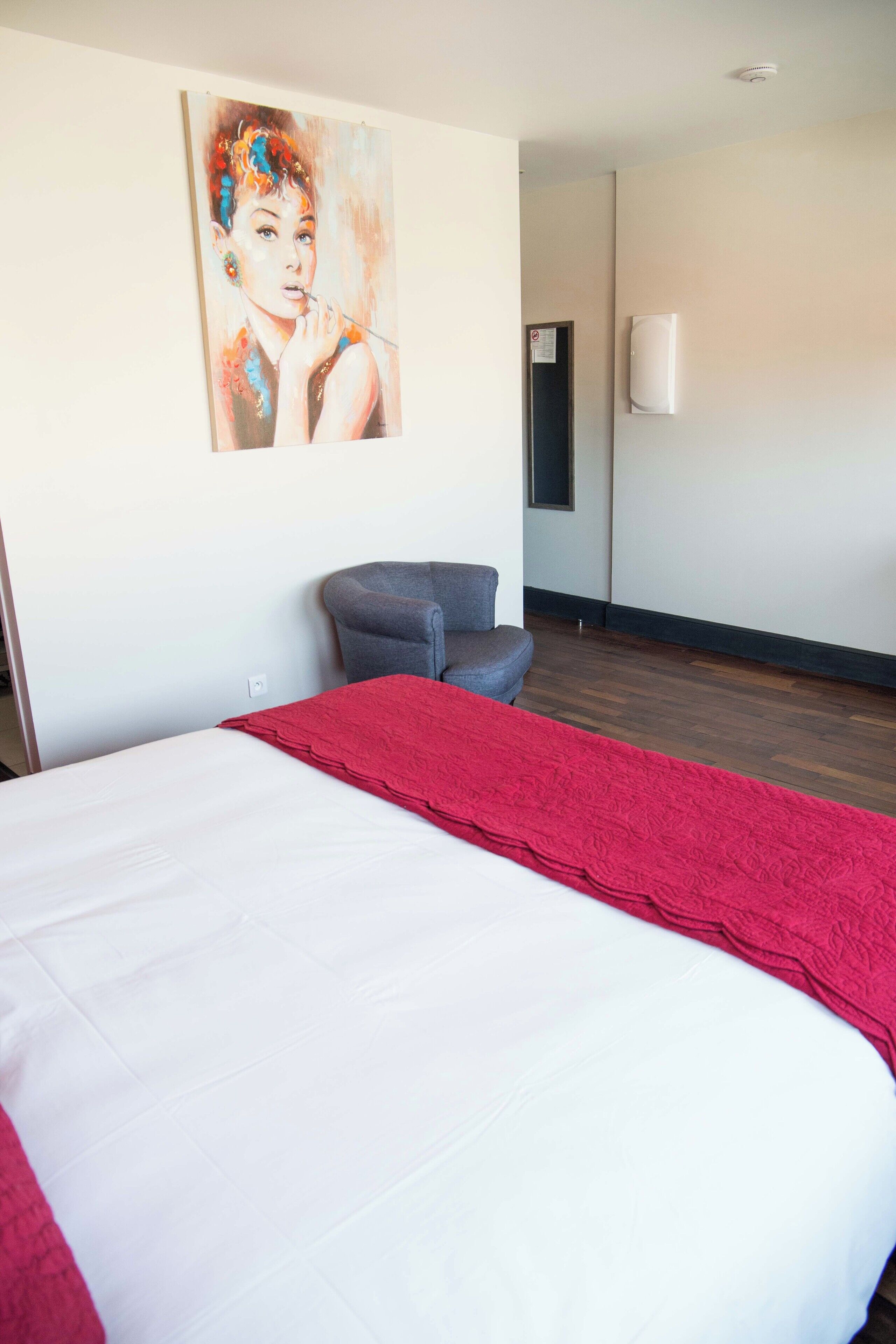 A hotel room with a large bed, a red throw blanket, a gray armchair, a painting on the wall, and a white air conditioning unit.