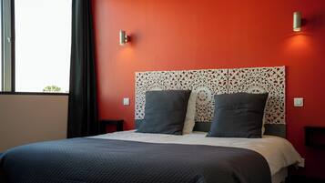 Quarto de hotel com parede vermelha, cabeceira estampada, duas mesinhas de cabeceira e cama com colcha escura.