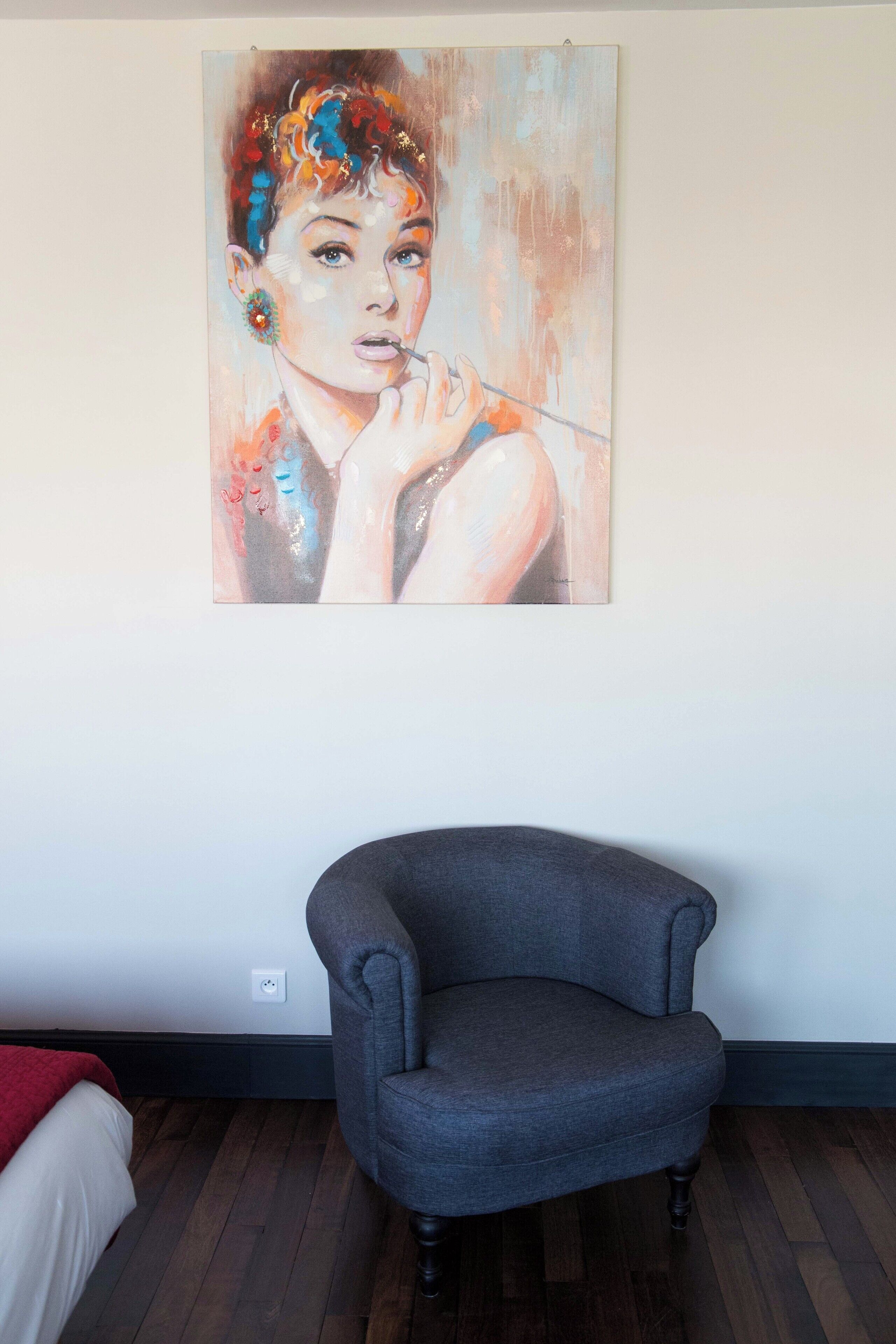 A room with a gray armchair, a painting on the wall, and a bed with a red blanket.