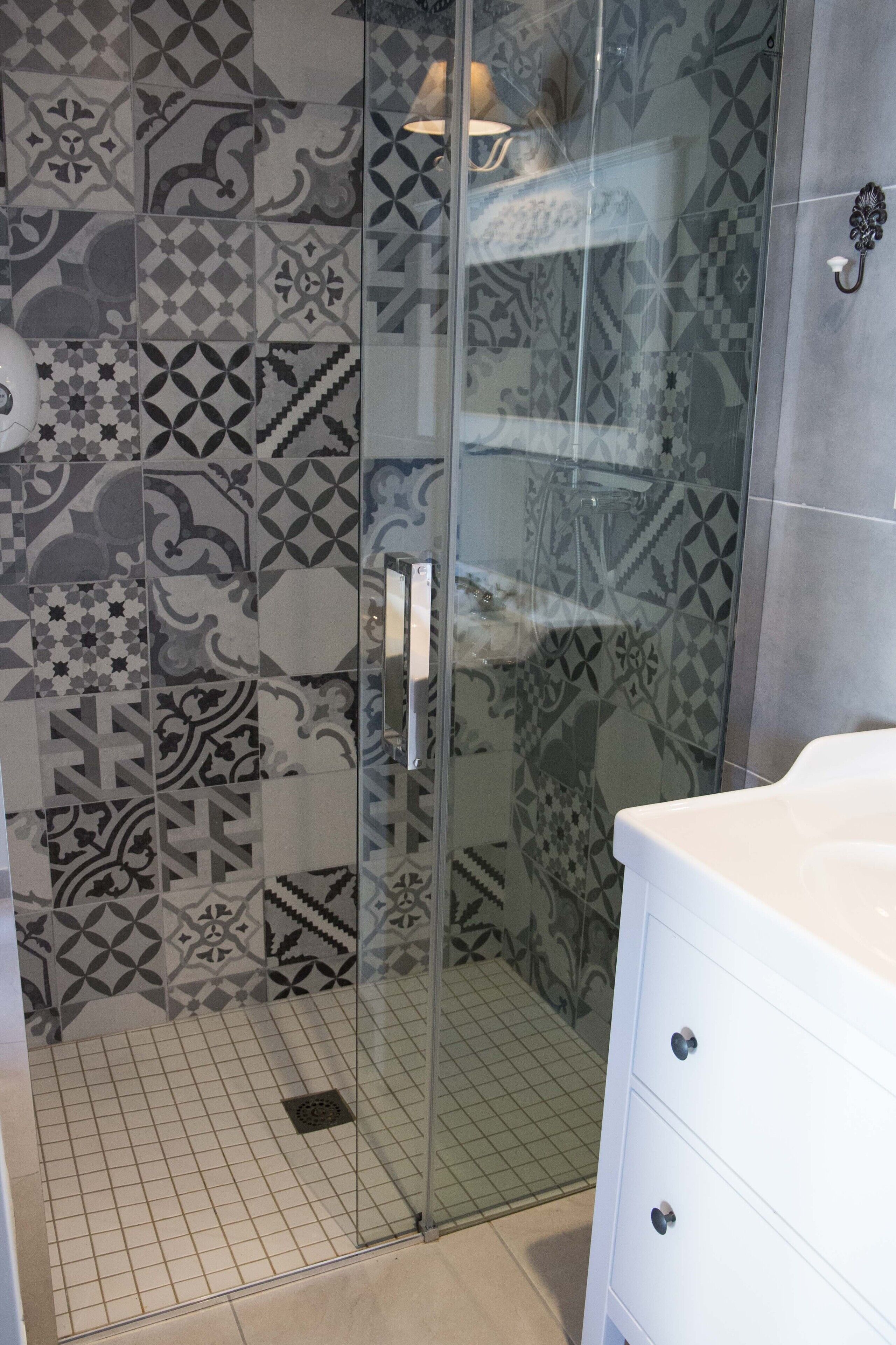 Un baño con baldosas estampadas, cabina de ducha de vidrio y tocador blanco con cajones.