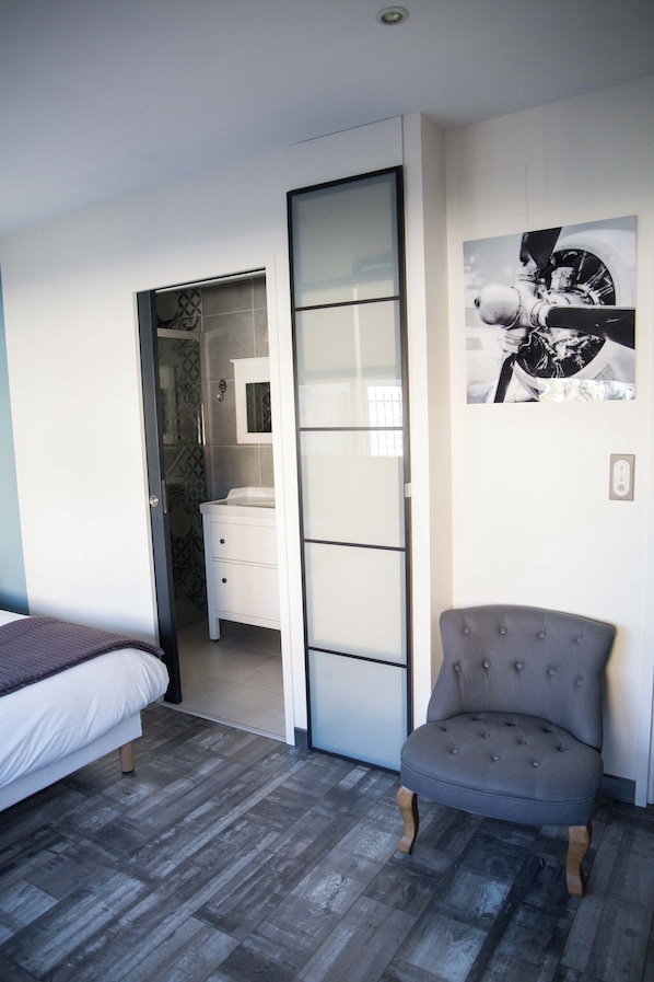 A bedroom with a bed, a grey upholstered chair, a white dresser, and a sliding door leading to a bathroom.