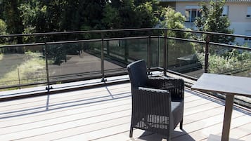 Un balcon avec une chaise en osier et une table, donnant sur un jardin et un immeuble.
