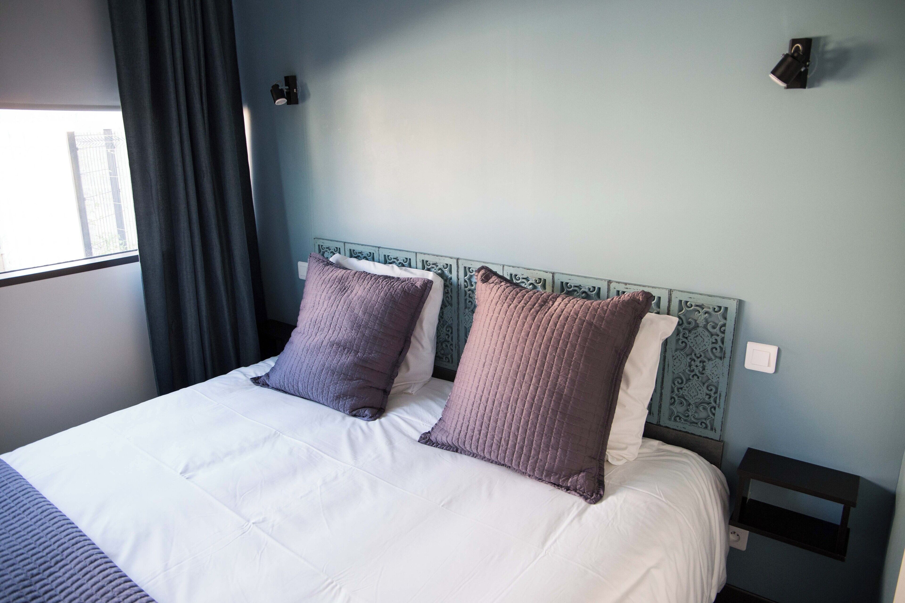 A neatly made bed with white and purple bedding, a blue headboard, and a small bedside table with a lamp.