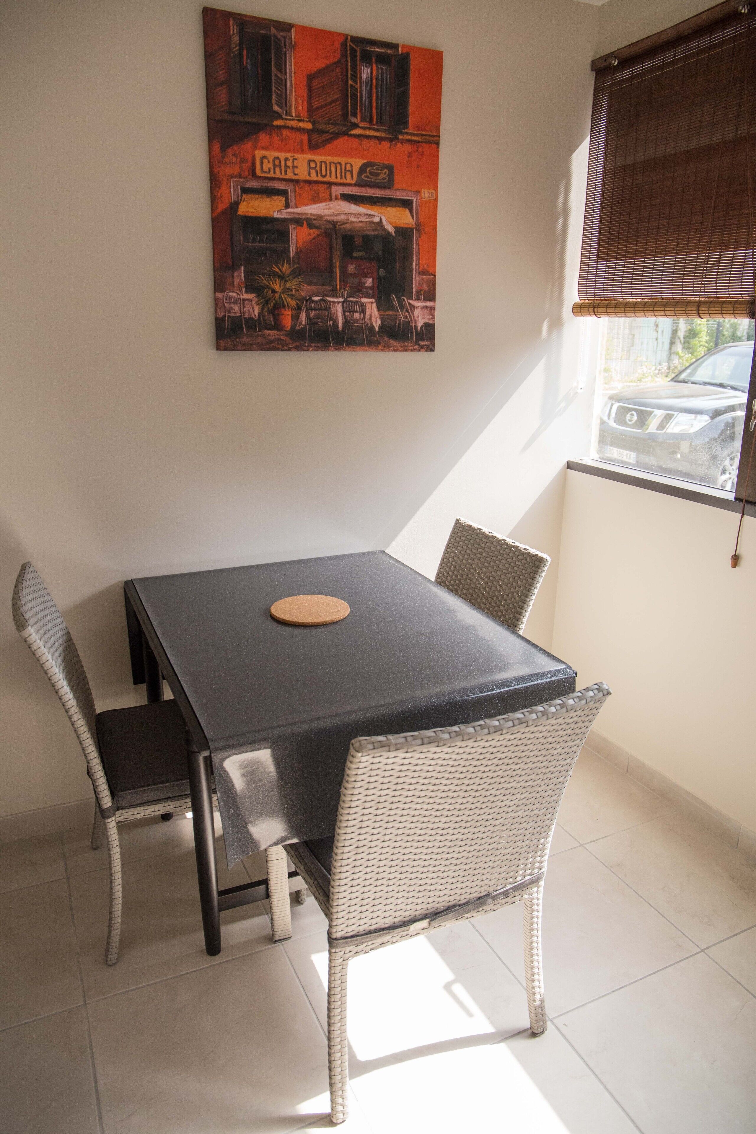 A dining area with a table and four chairs, a painting of a cafe on the wall, and a window with blinds.