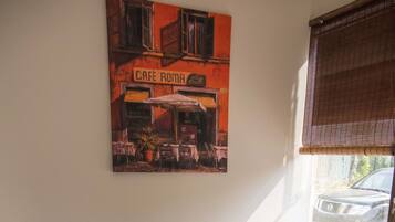 Uma área de refeições com uma mesa e quatro cadeiras, um quadro de um café na parede e uma janela com persianas.