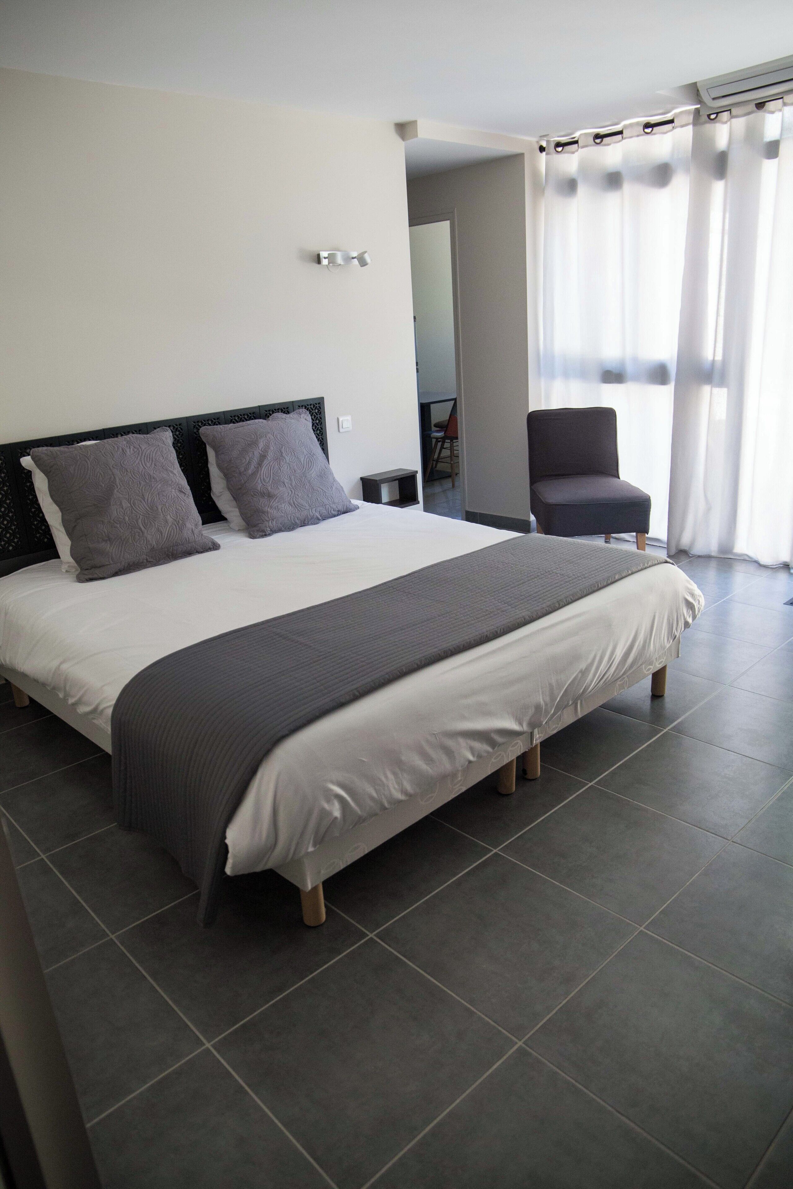 Ein modernes Hotelzimmer mit einem großen Bett, grauer Bettwäsche, einem dunklen Kopfteil und einem Sessel in der Nähe eines Fensters mit Vorhängen.