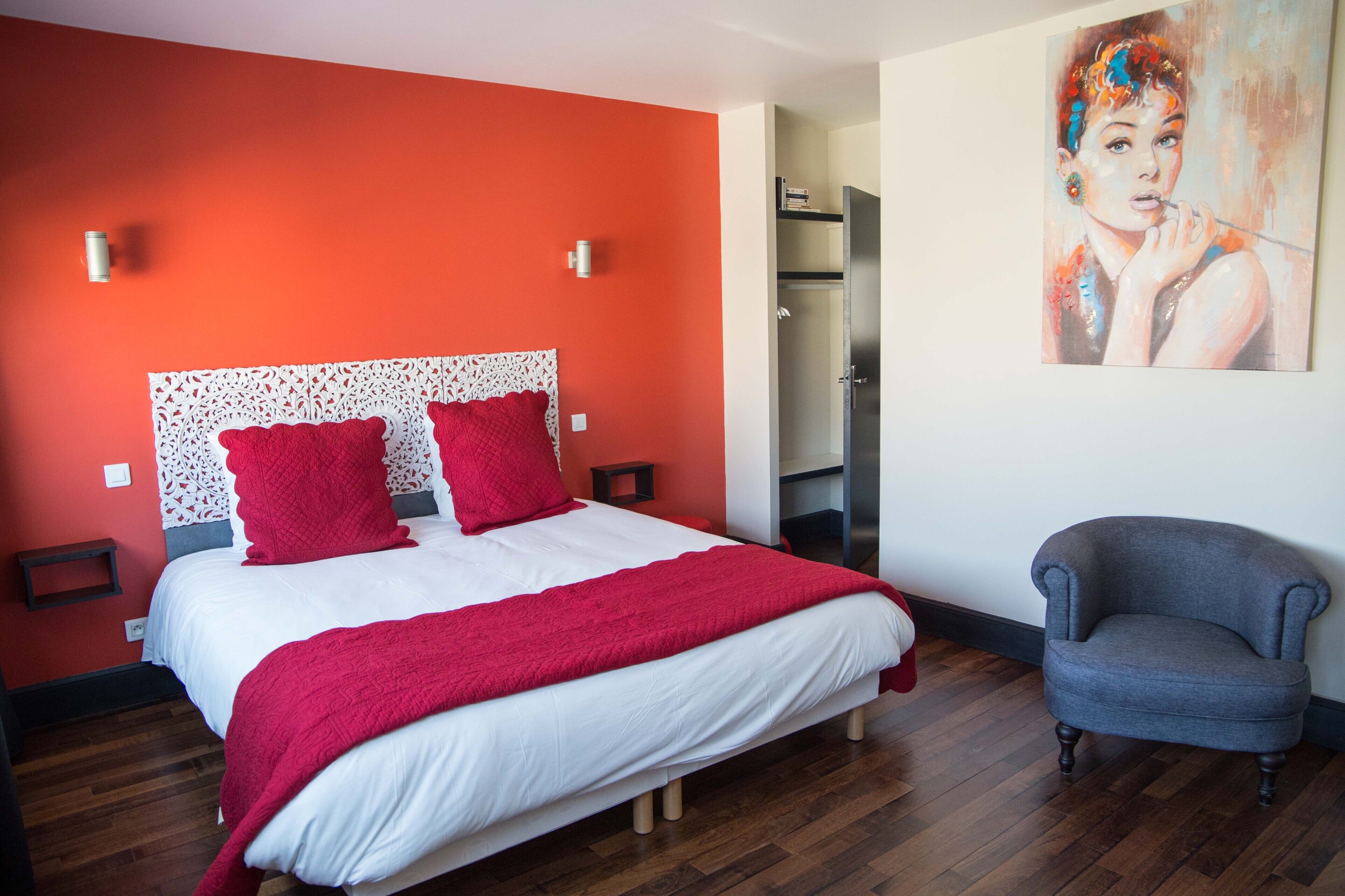 A bedroom with a bed, red pillows, a grey armchair, and a painting on the wall.