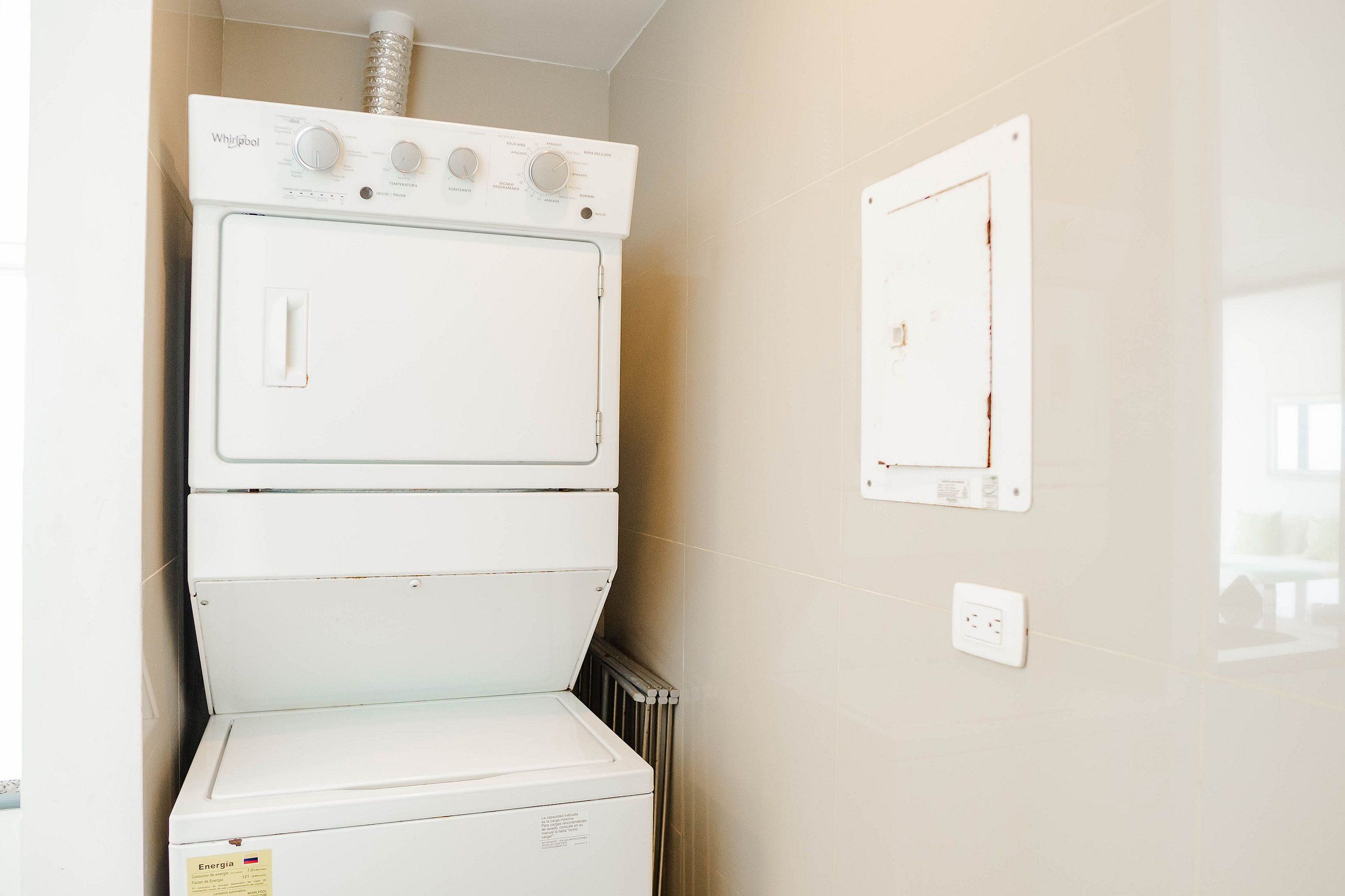 A stacked washer and dryer unit in a laundry room with a visible energy label and a wall-mounted electrical outlet.