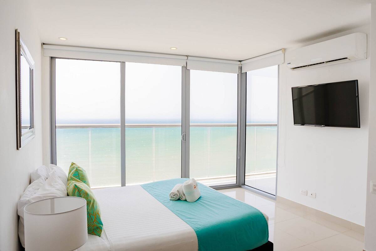 A hotel room with a bed, a TV, and a balcony with a view of the sea.