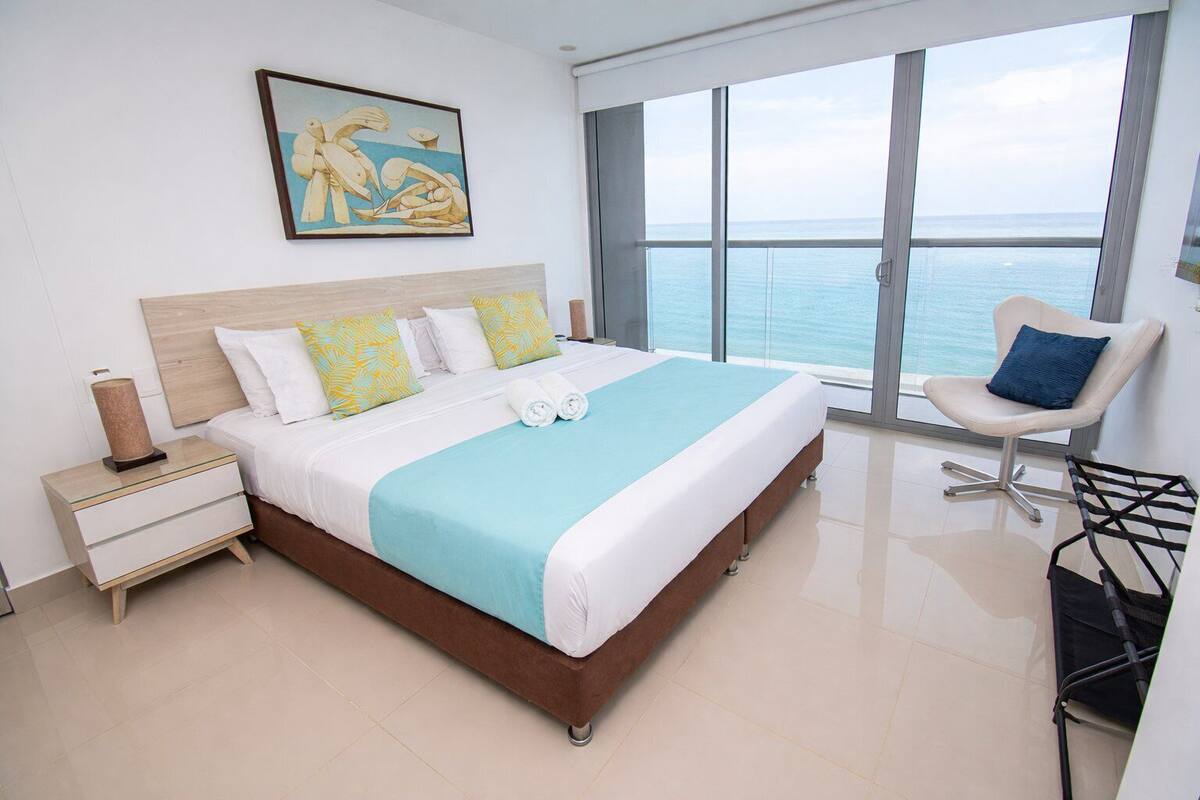 A modern hotel bedroom with a large bed, a nightstand, a chair, and a view of the sea.