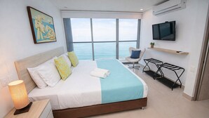 A modern hotel room with a large bed, a TV, a chair, and a view of the ocean.