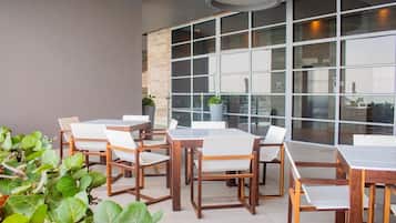 An outdoor seating area with wooden tables and chairs, surrounded by large windows and potted plants.