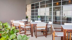 An outdoor seating area with wooden tables and chairs, surrounded by large windows and potted plants.