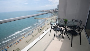 A balcony with a glass table and chairs, overlooking a beach and ocean.