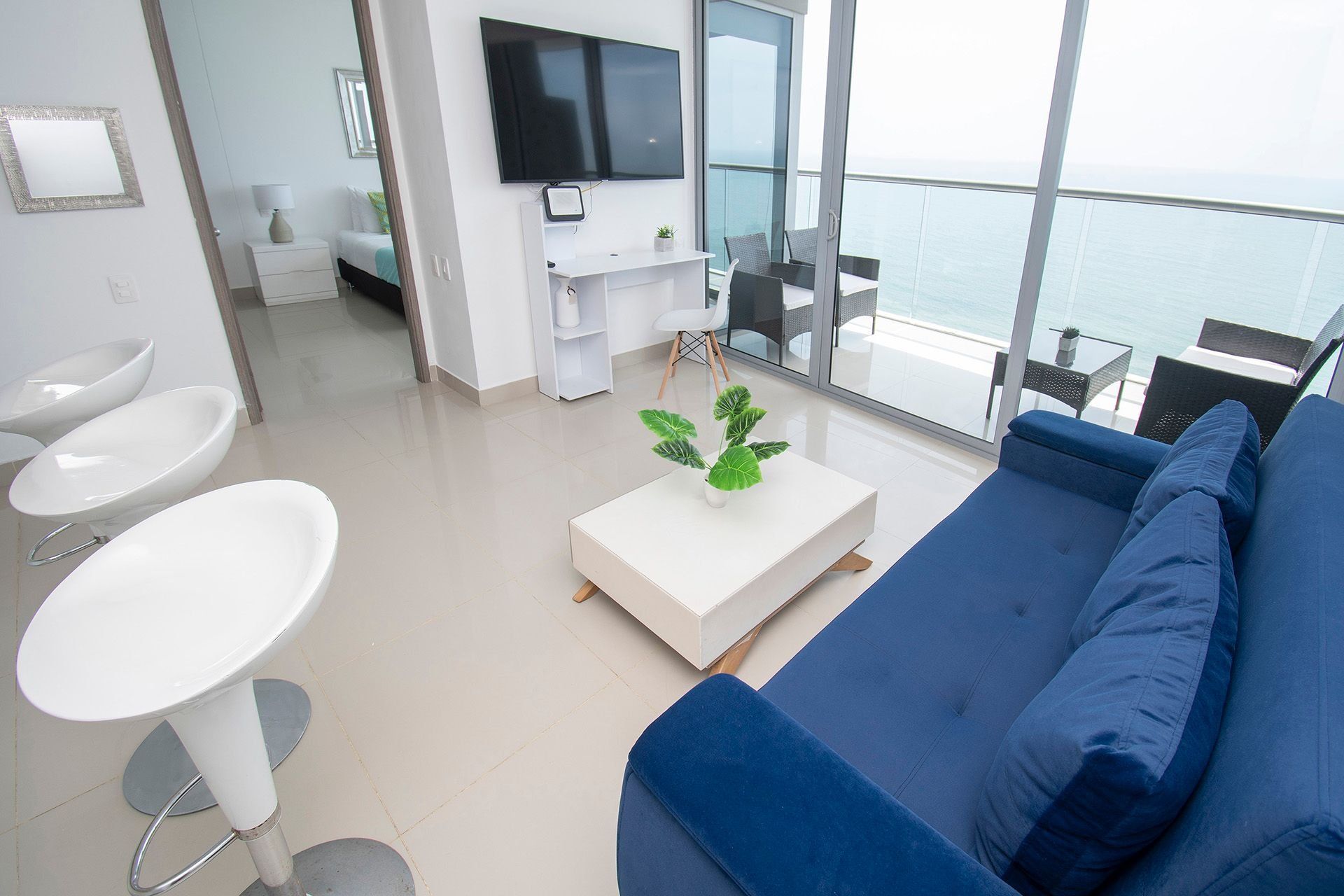 A modern living room with a blue sofa, white coffee table, and a TV mounted on the wall. There is a balcony with a view of the sea.
