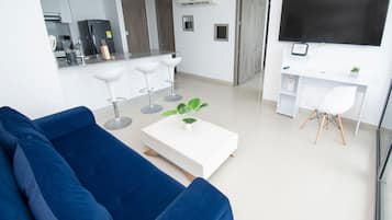 Un salon moderne avec un canapé bleu, une table basse blanche agrémentée d’une plante, un coin repas avec une table et des chaises blanches, et un téléviseur fixé au mur.