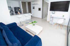 Una sala de estar moderna con un sofá azul, una mesa de centro blanca con una planta, un comedor con una mesa y sillas blancas, y un televisor montado en la pared.
