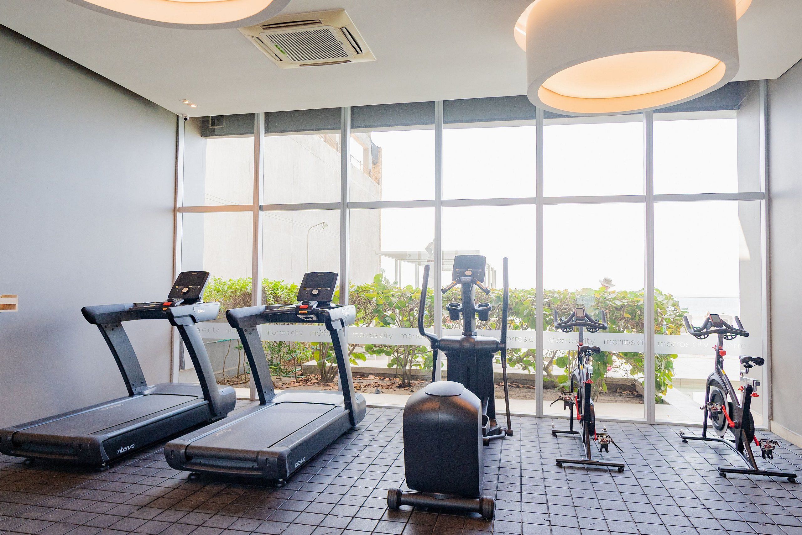 A gym with treadmills, an elliptical, and a stationary bike, large windows, and a tiled floor.