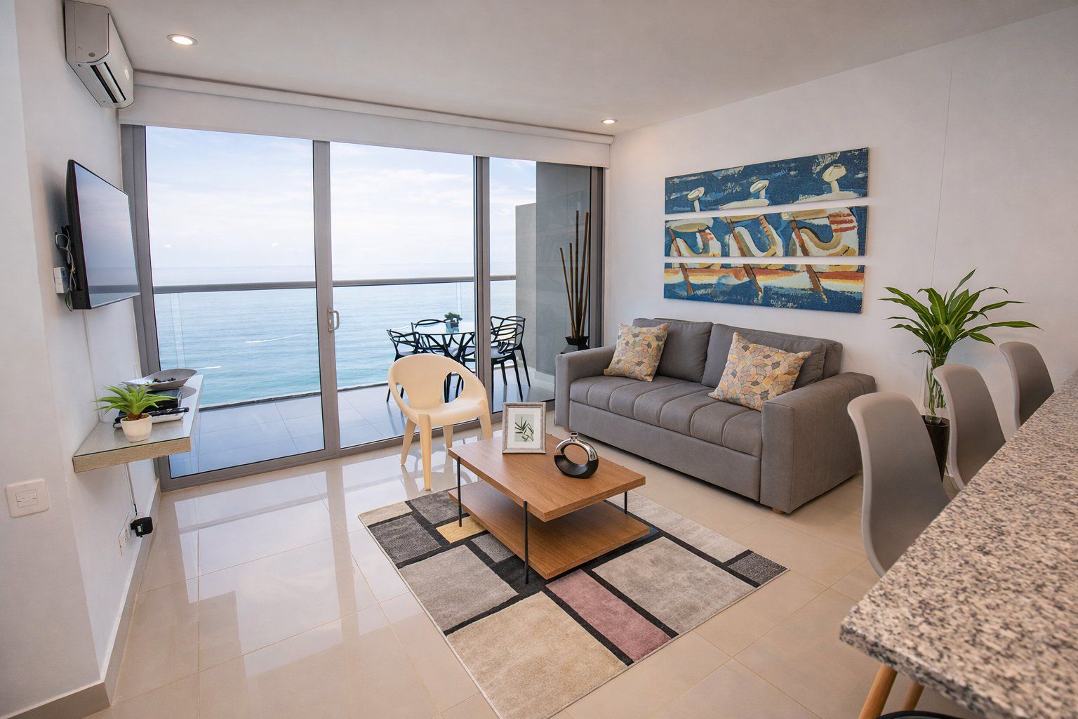 Un salon moderne avec un canapé gris, une table basse en bois et un téléviseur fixé au mur. Une grande fenêtre offre une vue sur la mer.