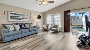 Un salon comprenant un canapé, deux fauteuils, un ventilateur de plafond, un tableau et une vue sur une piscine.