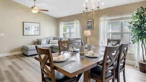 Un coin repas avec une table en bois dressée pour quatre personnes, entourée de chaises. On peut voir un ventilateur de plafond et un lustre au-dessus.