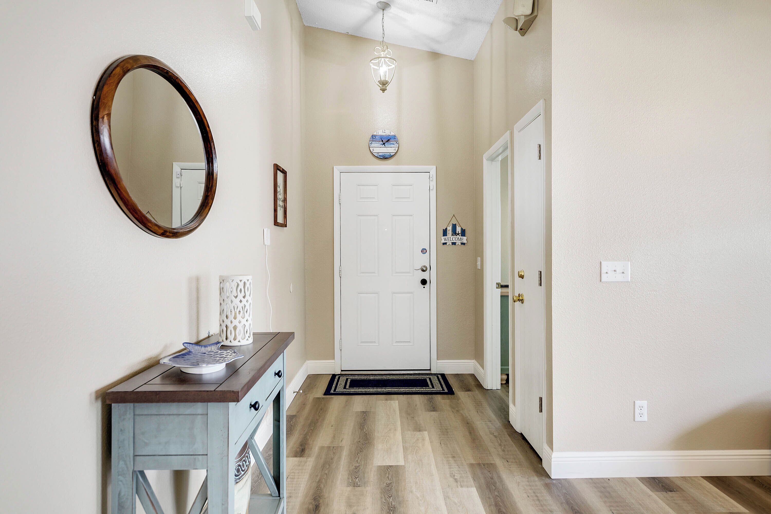 A hallway with a wooden floor, a white door, a round mirror, and a small table with a lamp.