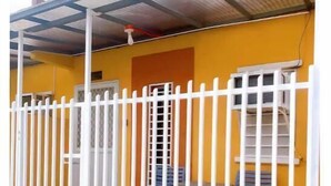 Un bâtiment jaune avec une clôture blanche, un porche couvert et une fenêtre avec des stores.