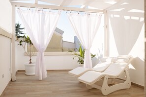 A sunlit room with a wooden floor, a white curtain, a potted plant, and a white lounge chair.
