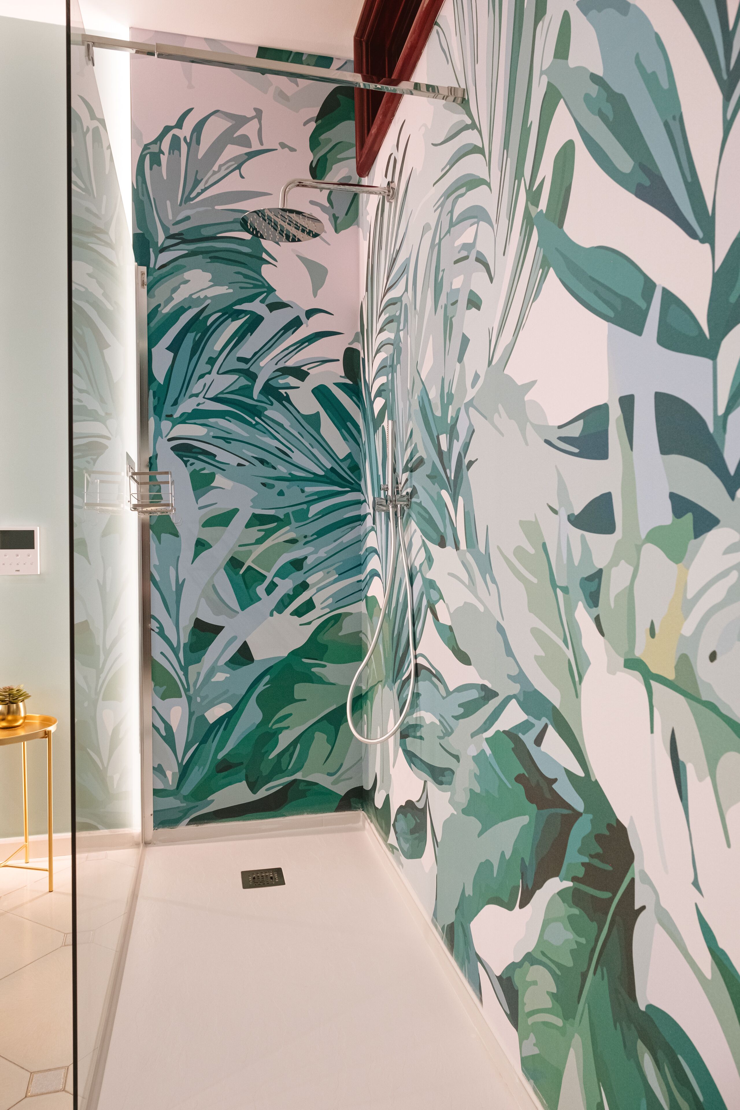 Une douche avec un motif de feuilles tropicales sur le mur, une porte en verre et une petite table sur laquelle est posée une plante.