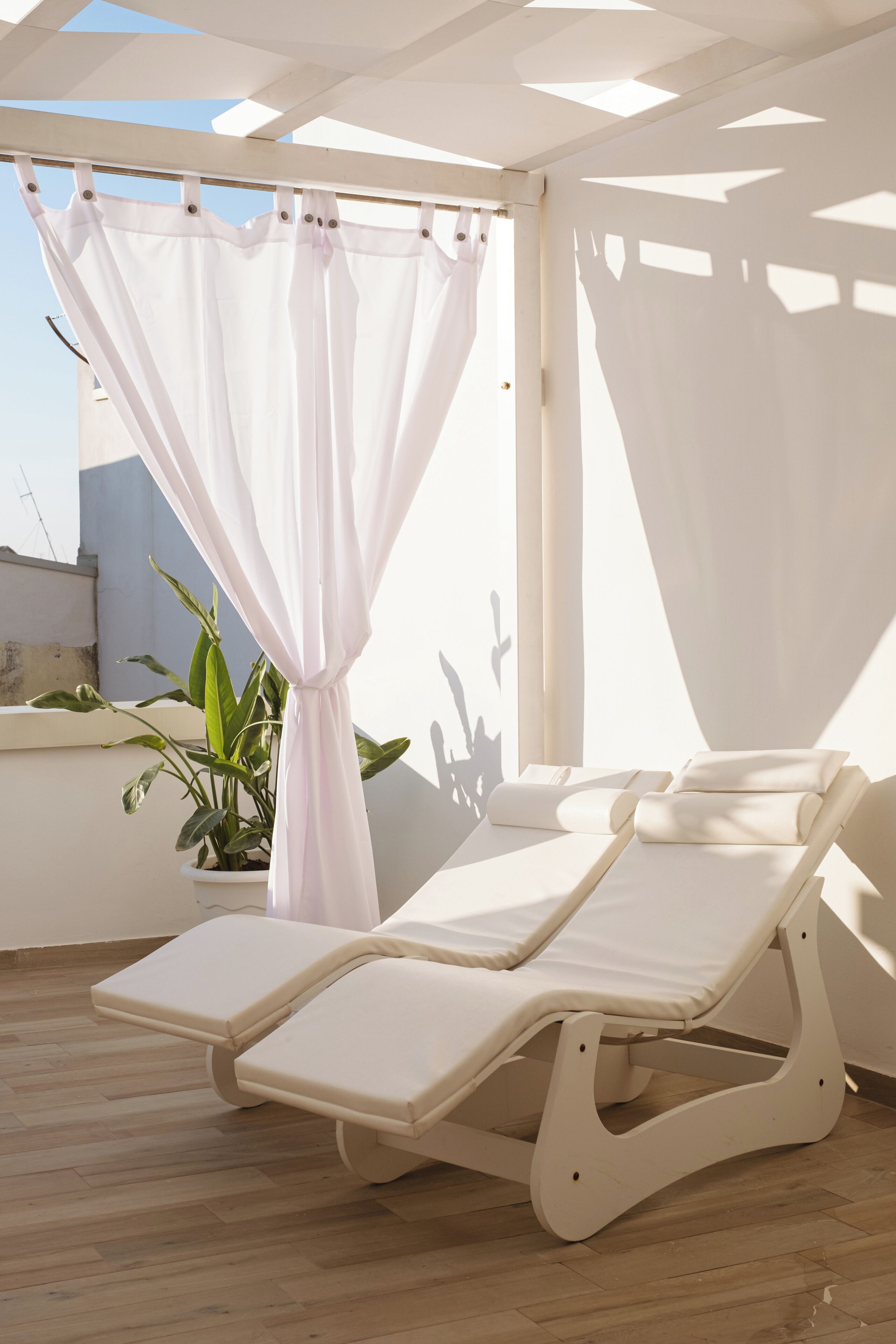 Una stanza luminosa con soffitto bianco, una pianta in vaso e un lettino prendisole bianco con cuscini.