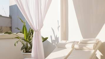 Una stanza luminosa con soffitto bianco, una pianta in vaso e un lettino prendisole bianco con cuscini.