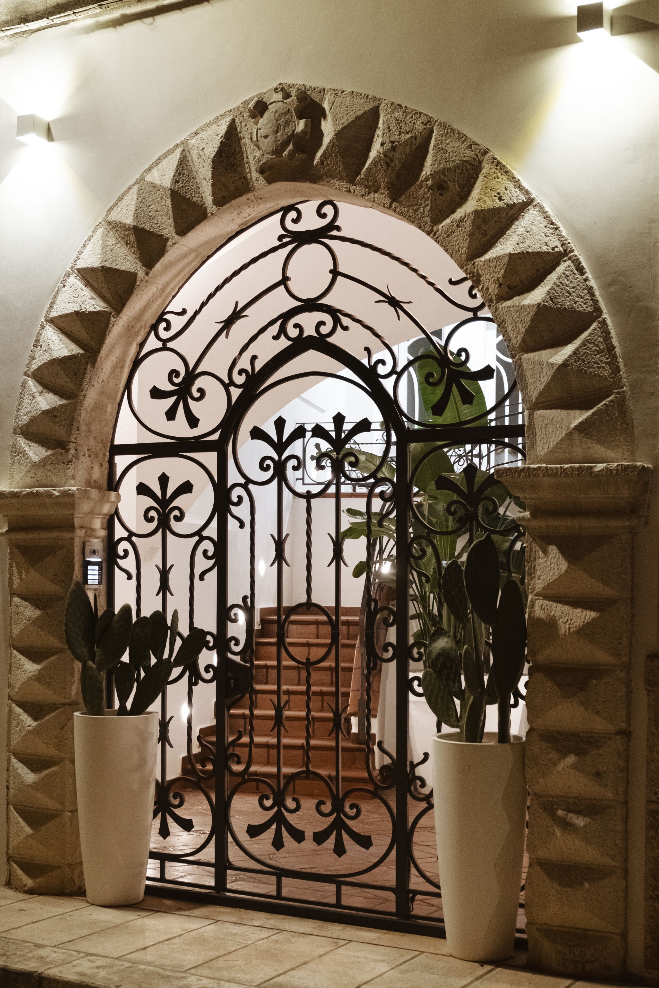Uma porta em arco com grade de ferro forjado, ladeada por dois vasos de plantas, dando acesso a uma escadaria.