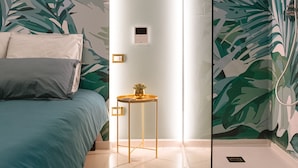 Un dormitorio con cama, una mesita de noche, una ducha con puerta de vidrio y una pared con estampado de hojas.