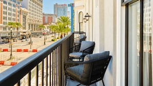 Un balcon avec deux fauteuils en osier noir et une table, donnant sur une rue bordée de palmiers et d’immeubles.