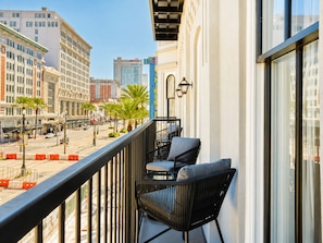 Ein Balkon mit zwei schwarzen Korbstühlen und einem Tisch, mit Blick auf eine Straße mit Palmen und Gebäuden.