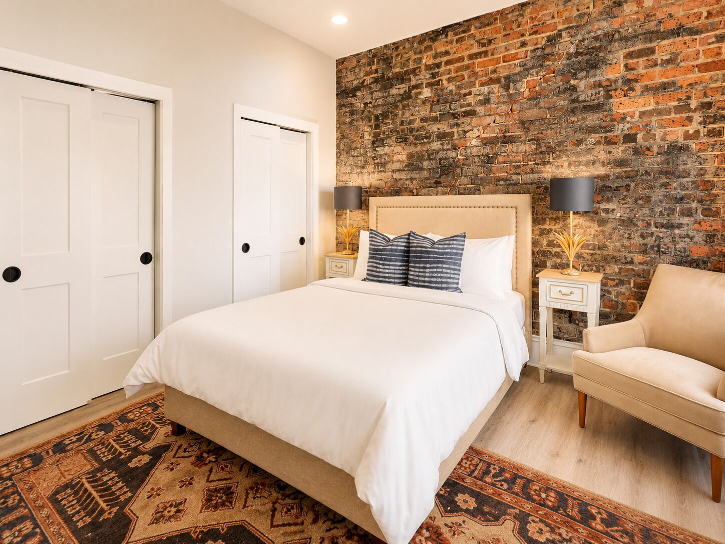 A bedroom with a brick wall, a bed with white bedding, two bedside tables with lamps, a chair, and a patterned rug.