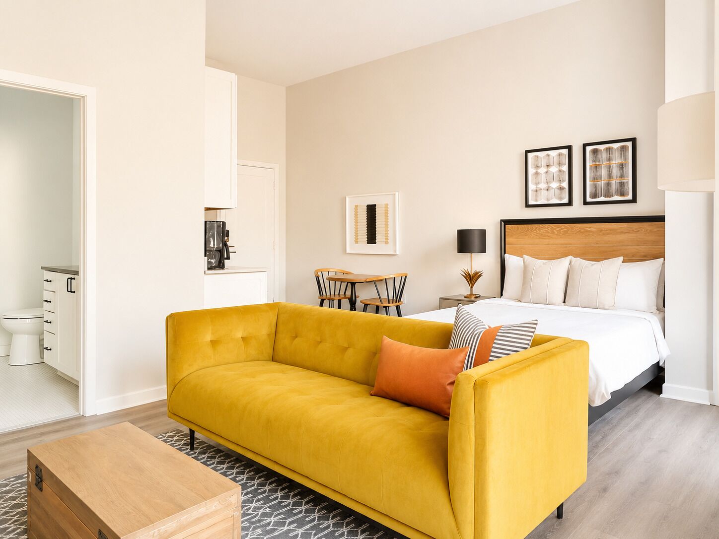 A hotel room with a yellow sofa, a bed with white bedding, a wooden headboard, and two framed artworks on the wall.