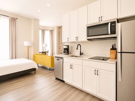 A modern hotel room with a bed, a yellow sofa, a kitchen area with a microwave, sink, and refrigerator.