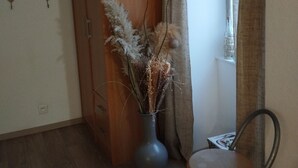 Une pièce avec une armoire en bois, un vase contenant des plantes séchées, une chaise et une fenêtre avec des rideaux.