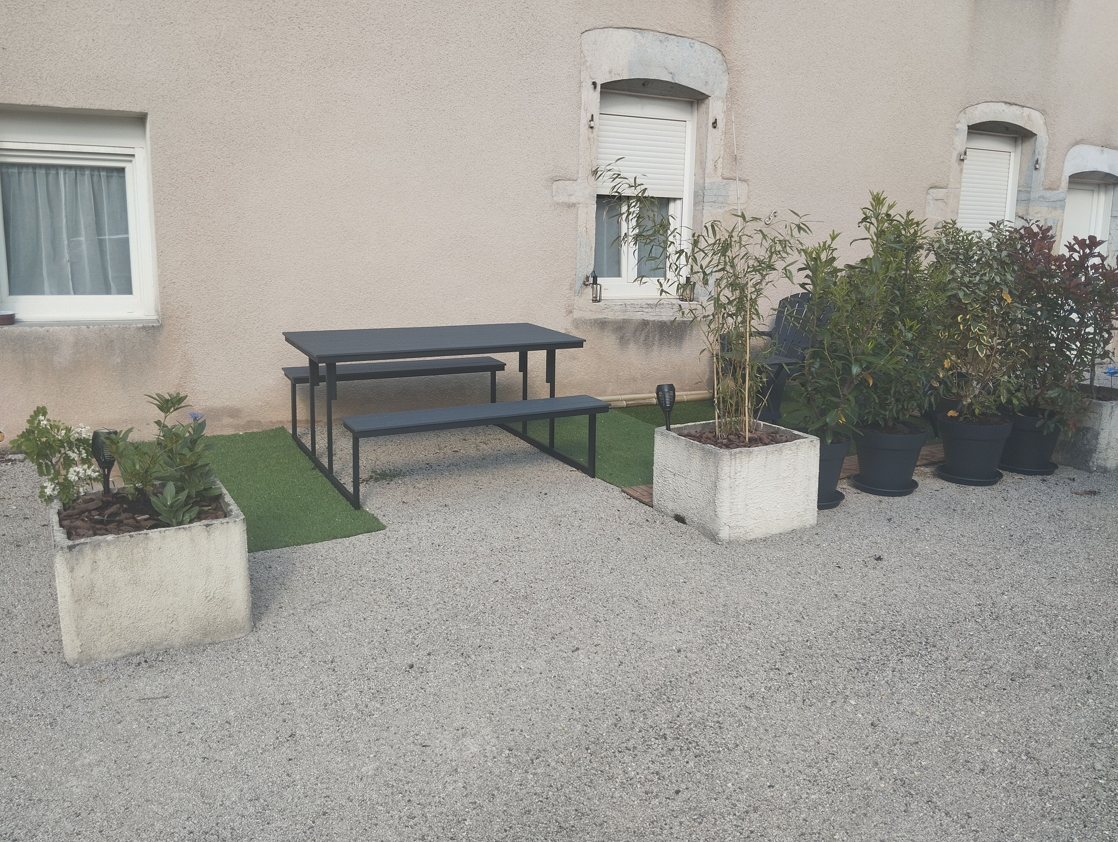 Een terras met een tafel en bankjes, omringd door potplanten en een klein gazon.