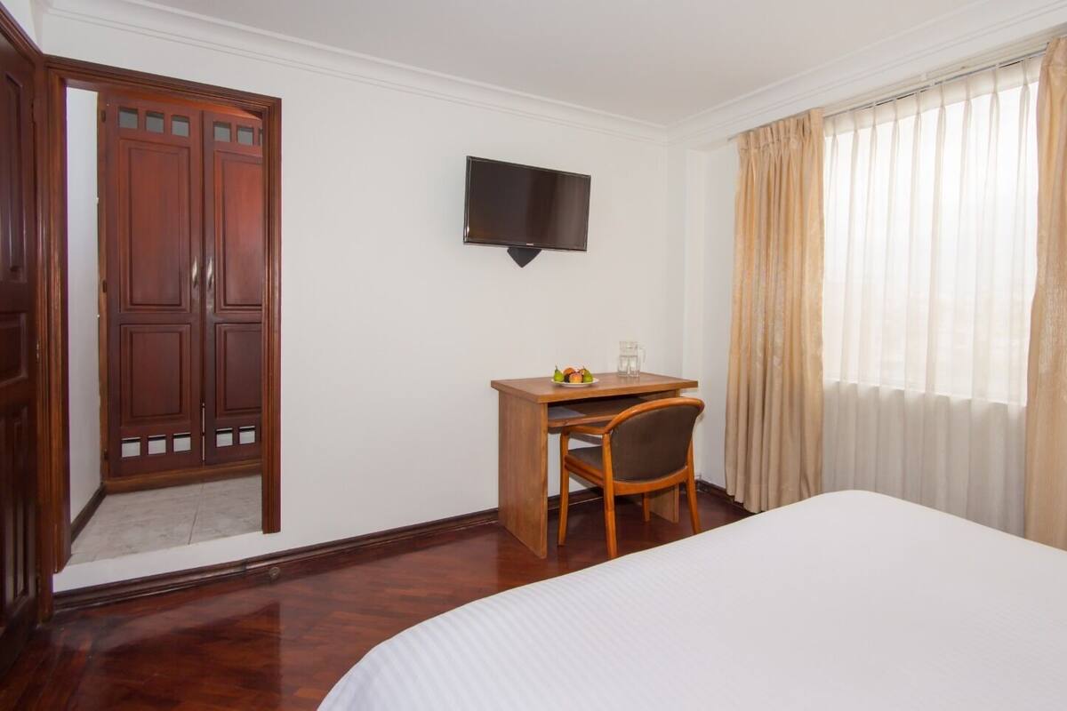 Quarto de hotel com cama, escrivaninha com cadeira, televisão na parede e janela com cortinas.