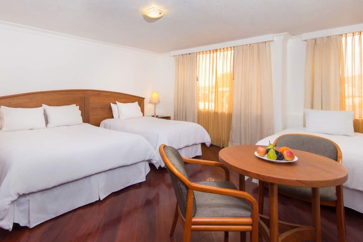 Quarto de hotel com duas camas individuais, cabeceira de madeira, uma mesinha redonda com uma cesta de frutas e uma cadeira.