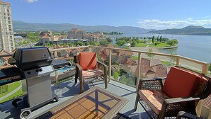 Un balcón con muebles de mimbre, una parrilla y vistas a un lago y edificios.