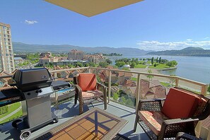 En balkon med fletmøbler, grill og udsigt over sø og bygninger.